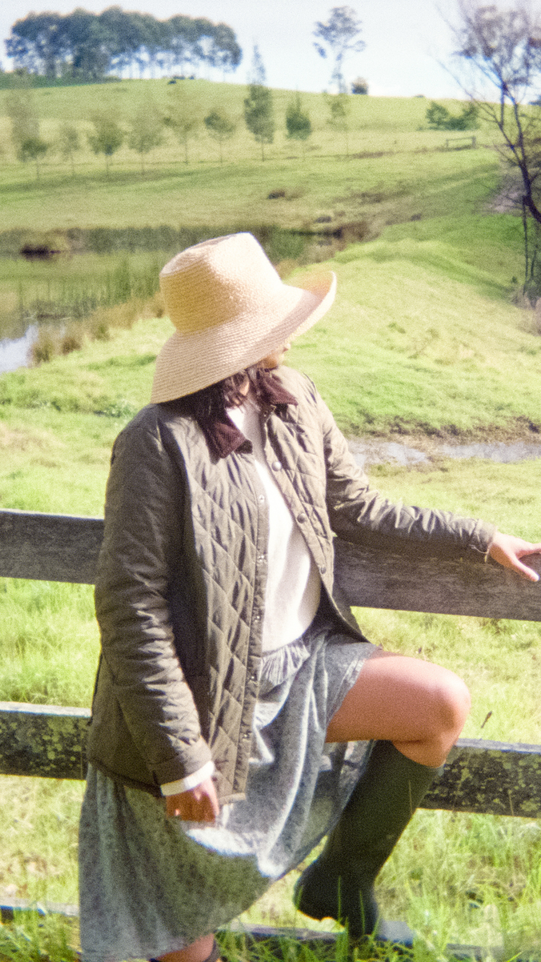 Woman sitting on a wooden fence in a pastoral setting wearing chic clothing and raffia hat 