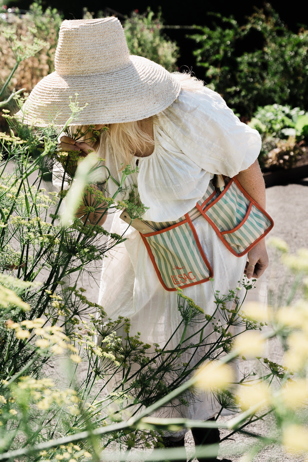 Person wearing a wide-brimmed hat and white dress with striped garden belt le sac  in a garden setting.