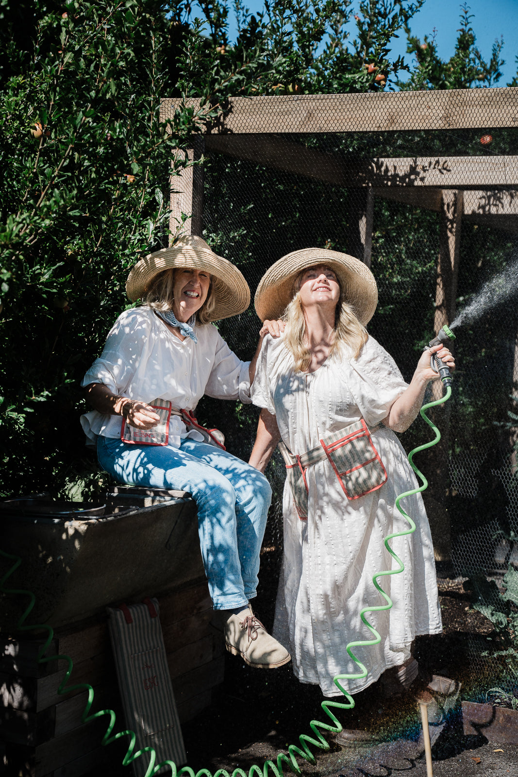 Two women in vintage clothing with hats, one sitting and one standing with a hose, outdoors. Wearing raffia garden hats 